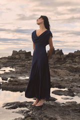 Caroline Cowl Satin Dress in Midnight, side view worn on rocks by the sea